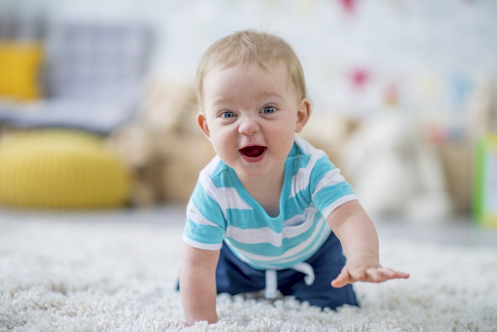 A Caucasian baby boy is crawling towards the camera while laughing. He is giggling and looking at the camera. He is at a daycare center.
